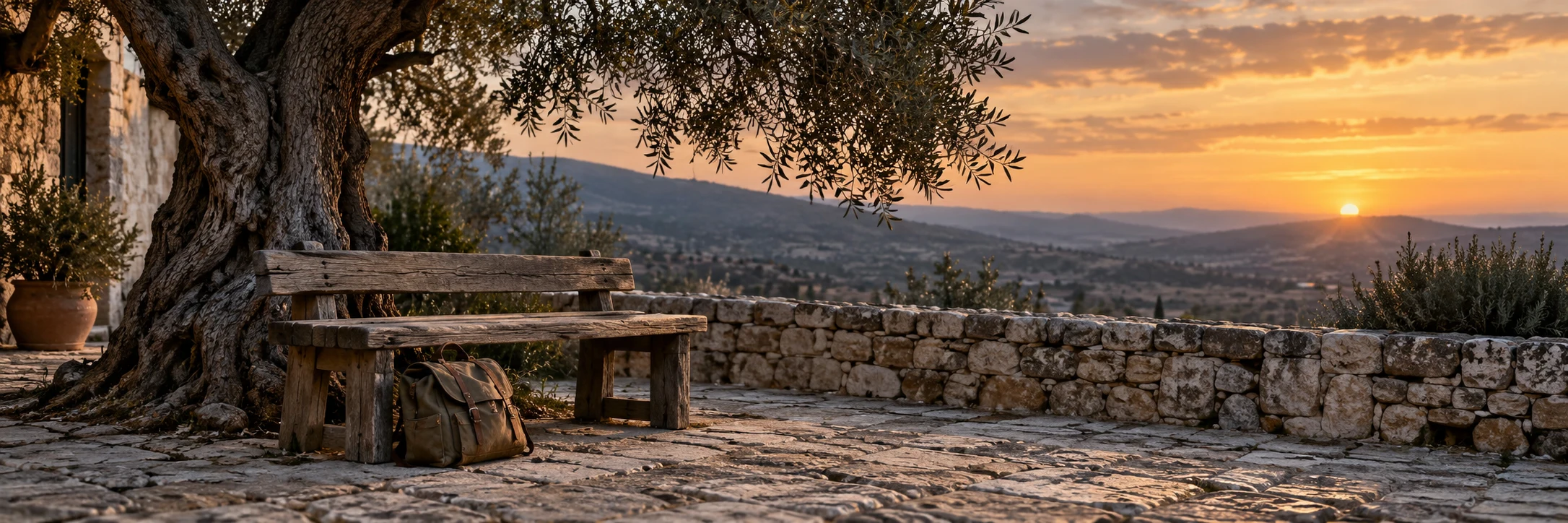 A canvas travel bag rests beside a weathered wooden bench beneath an olive tree in a quiet stone courtyard at dusk.