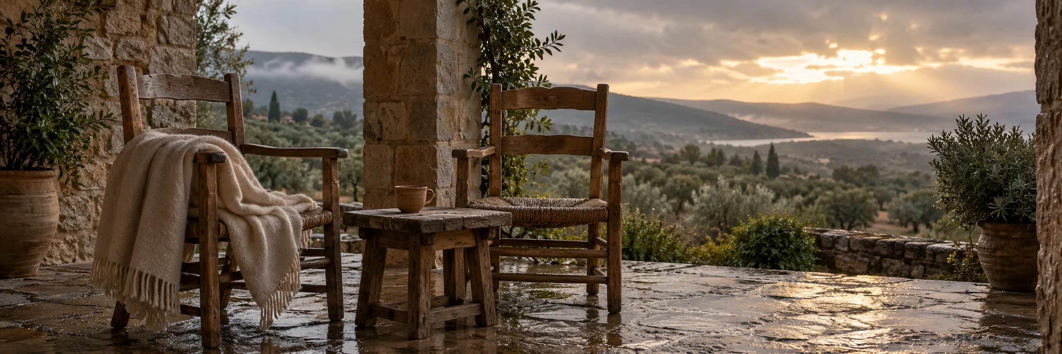 Two wooden chairs, a wool blanket and a cup sit on a sheltered stone porch overlooking quiet countryside after rain.