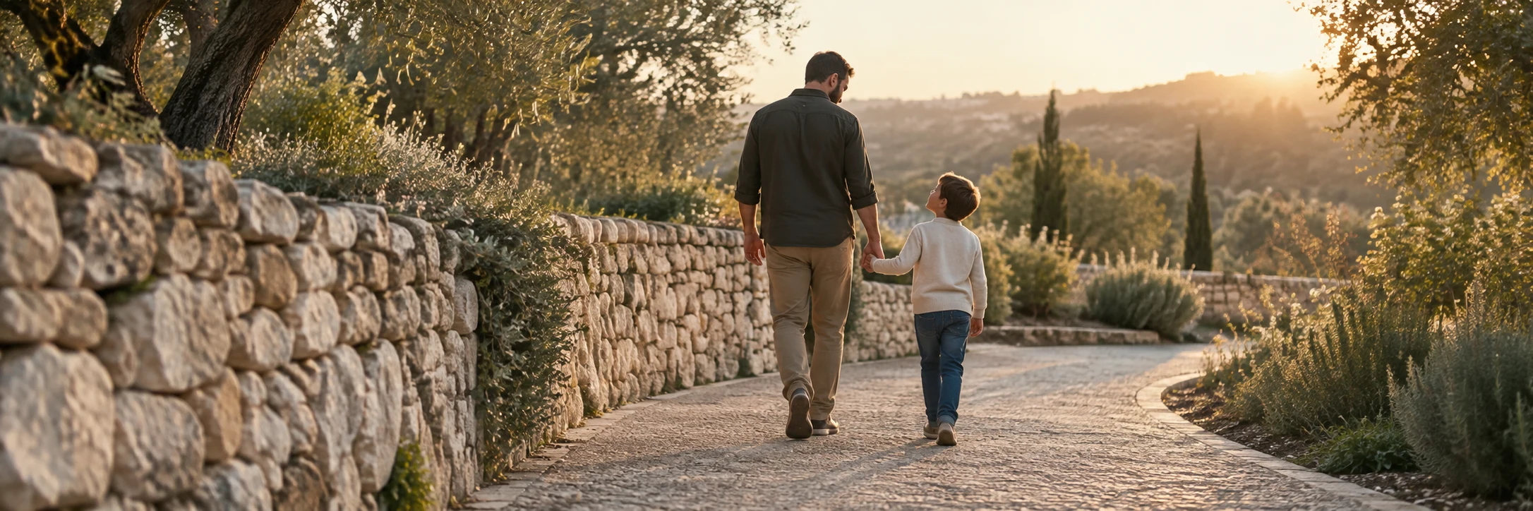 A father and child hold hands while walking beside a weathered stone wall and olive-green foliage in warm late-afternoon light.